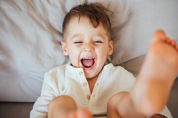 Young boy with light brown hair is joyfully laughing while lying on a soft gray couch, showcasing a...