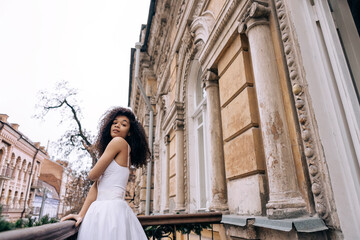 Obraz premium A young African woman in an elegant white dress and jewelry stands on the balcony of a retro house