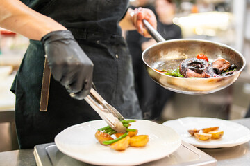 Chef plating gourmet vegetables in a professional kitchen. Black gloves, tongs, and frying pan suggest precision, culinary artistry, and fine dining preparation.