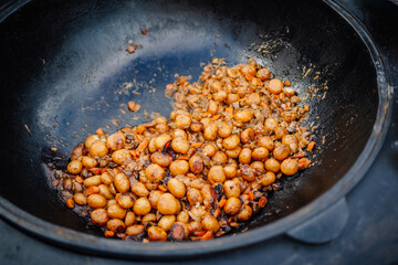 Close-up of roasted baby potatoes with onions and carrots cooking in a large black cast iron pan, possibly at an outdoor event or rustic gathering.