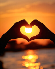 Fototapeta premium Silhouetted hands form a heart shape against a sunset backdrop, with the sun glowing in the center, creating a warm and romantic atmosphere.