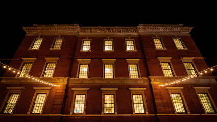 A three-story brick building is photographed at night, its many symmetrical windows glowing warmly. Festive strings of lights cross the foreground, accentuating the inviting facade.
