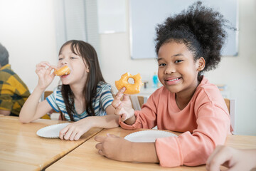 Happy group of diverse school children black girl eating donuts together at desks with their teacher nearby, celebrating a fun classroom break.
