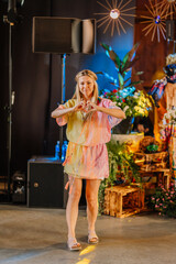 Smiling woman making a heart gesture with hands at an indoor Hippie festival. She wears a colorful boho dress and headband, standing in a vibrant decorated space.