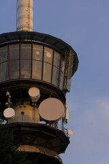 A vertical shot captures the top of a modern communication tower, with its distinctive circular windows and multiple white satellite dishes against a clear blue sky.