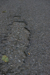 A cracked and weathered asphalt road with visible gravel and a single fallen leaf, creating a textured and moody image of urban decay, damage, and neglect.