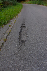 A cracked and weathered asphalt road with visible gravel and a single fallen leaf, creating a textured and moody image of urban decay, damage, and neglect.