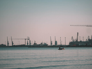 Obraz premium Tranquil harbor photo of a small boat on calm waters with a distant industrial port and cranes under a soft sky. Perfect for travel decor, maritime art, and eco-friendly designs.