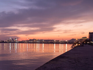 Fototapeta premium Breathtaking sunset photo of a city skyline reflecting on calm waters, with a serene promenade in the foreground. Ideal for travel decor, coastal art, and eco-friendly designs.