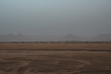 Dusty Sahara desert landscape in Morocco with flat barren ground and distant mountain silhouettes fading in haze under overcast sky atmospheric view