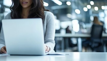 A woman with long dark hair is sitting at a table, using a silver laptop in a brightly lit office.