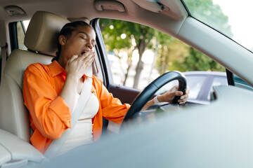 Tired African American woman yawning while driving car with seatbelt. Concept of fatigue, daily commuting stress, and urban lifestyle challenges.