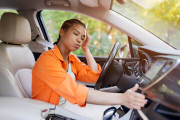Tired African American woman sitting in car, wiping forehead and adjusting dashboard controls. Concept of summer heat, travel fatigue, and lifestyle