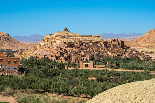 Ait Benhaddou kasbah in Morocco with ancient adobe houses built on hillside surrounded by palm trees desert landscape and Atlas Mountains  - Powered by Adobe