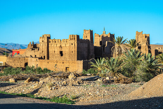 Ancient kasbah fortress in Morocco with tall adobe walls palm trees and rocky ground in foreground under clear blue sky and Atlas Mountains behind