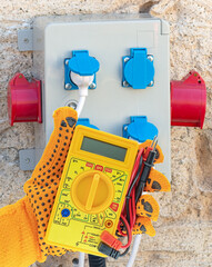 gloved hand with a multimeter on the background of an electric s