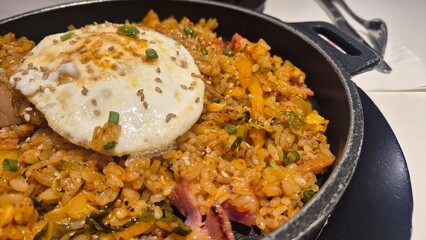 Close-up of Korean kimchi fried rice served in a hot pan with a fried egg on top, garnished with sesame seeds and green onions, traditional Asian comfort food
