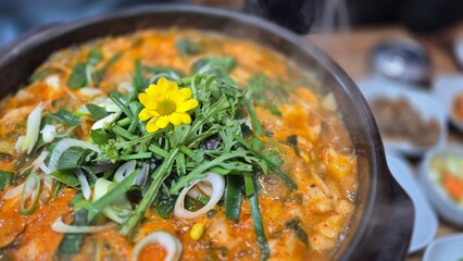 Korean spicy stew served in a hot pot with vegetables, kimchi, and broth, garnished with fresh greens and edible flower, traditional Asian comfort food.