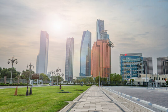 Fototapeta Abu Dhabi, UAE – August 16, 2025: Skyscrapers overlook a palm-filled park with fountains at sunset in the capital of the United Arab Emirates.