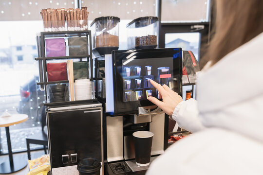 A woman in a white jacket buys a coffee drink or tea from a coffee machine at a gas station or store, selecting the desired drink on the screen with her finger.Close-up view from behind.