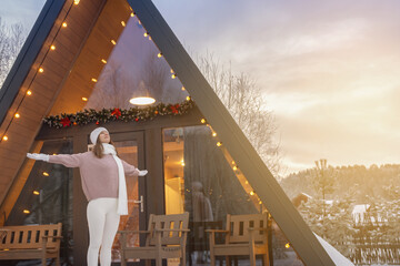 A woman rejoices at the sun and a new day on the threshold of her wooden house in nature, spreading her arms wide. new year and a new day in winter.