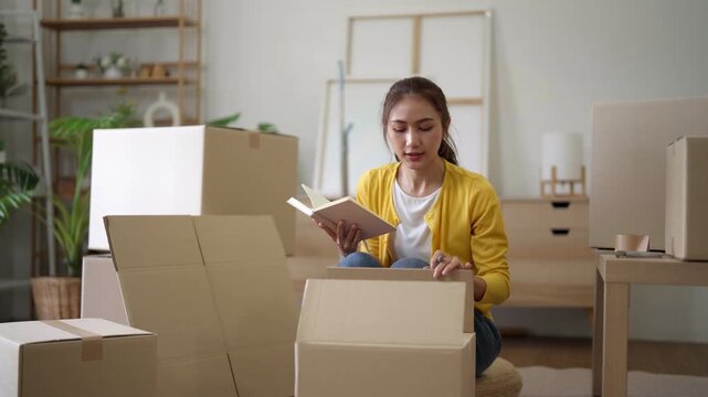 Asian adult woman organizes move boxes packs new home plans relocation settles personal items modern apartment living room