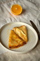 Delicious crepes suzette with orange syrup and fruit slices on white plate on white tablecloth, top view.
