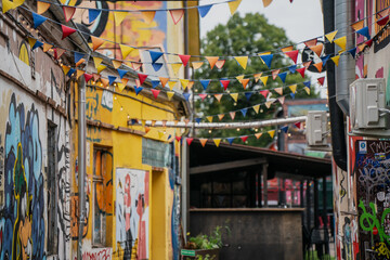 Fototapeta premium Urban alley decorated with colorful triangular pennant flags hanging between graffiti walls and murals, outdoor street scene featuring creative artistic expression and festive atmosphere