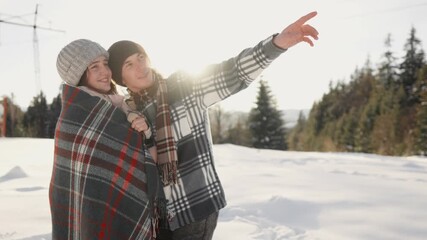 A joyful couple enjoys a beautiful snowy landscape, cozily wrapped under warm blankets. They happily point at the distant horizon, capturing a memorable moment filled with love and adventure together