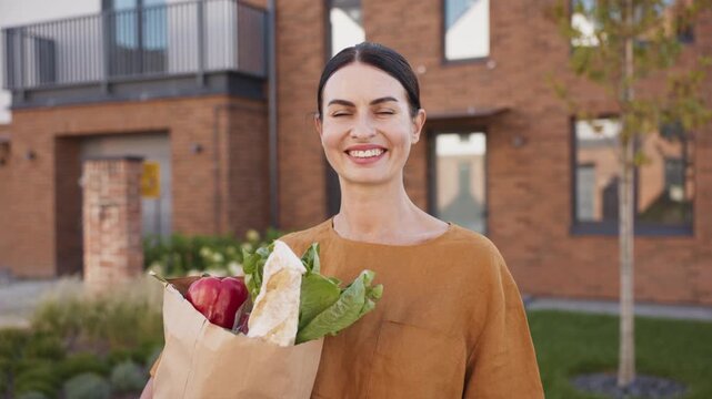 Close up portrait of smiling Caucasian woman holding grocery bag with bread and vegetables. Standing outdoors near modern brick residential house looking directly at camera with warm smile.