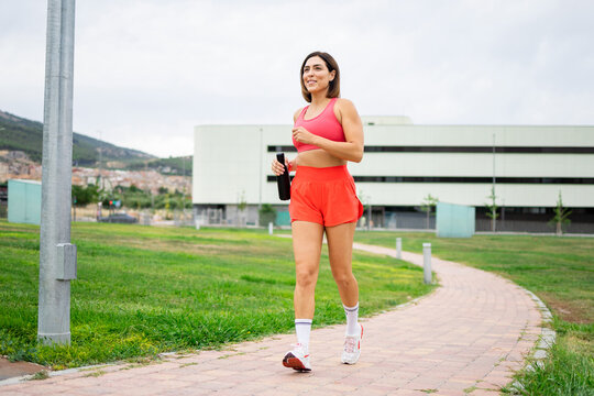 An attractive woman between 40 and 50 years old is exercising outdoors on a cloudy day. The girl is running with a water bottle. Concept of adult women training.