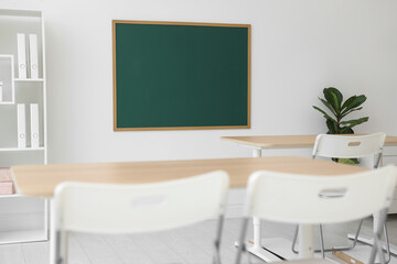 Fototapeta premium Blank green chalkboard, desks, chairs and plant in classroom
