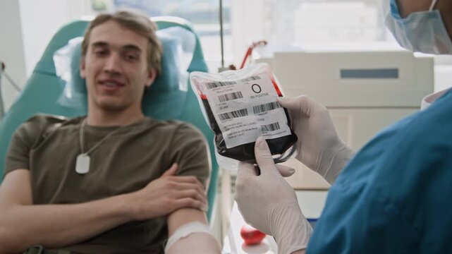 Over the shoulder shot of masked nurse checking blood bag and talking to young male donor, lying in chair in clinic