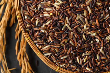 Top view of organic mixed rice grains in a bamboo basket with golden rice ears  on black stone