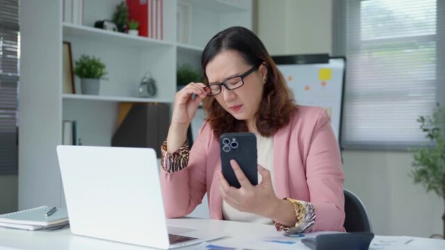 Professional businesswoman experiencing intense eyestrain, removing glasses while working on smartphone at modern office workspace, displaying signs of visual discomfort and work related stress