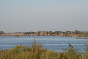 Calm water reflects the clear sky while lush greenery borders the lake. Nearby, power lines stretch across the horizon, adding a touch of industry to the serene nature