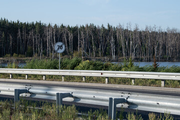 A speed limit sign indicates 70 kilometers per hour along a peaceful road. The setting features a serene lake bordered by trees and greenery, creating a tranquil view
