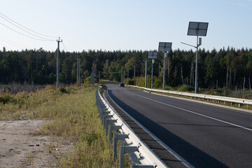 A long road stretches ahead, bordered by lush green trees and solar panels. The sunny day highlights the serenity of the rural landscape, blending nature with modern technology