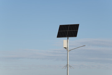 A solar panel is mounted on a tall metal pole, positioned in an open area under a bright blue sky. The environment is calm and sunny, highlighting renewable energy equipment