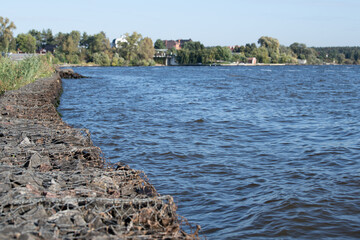 A peaceful riverbank extends along the water, featuring gentle waves and vibrant greenery. A small town is visible in the background under clear blue skies