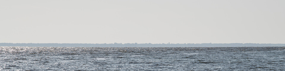 A serene ocean stretches towards a distant shoreline under a bright sky. The water sparkles in the sunlight, creating a peaceful atmosphere typical of a midday scene