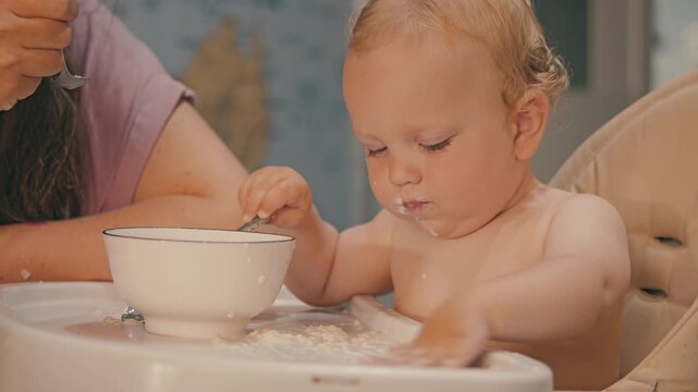 The baby is sitting in a high chair. The whole table is dirty with porridge. The parent is feeding the son with a spoon. The child takes the porridge from the bowl with his hand and tries to eat