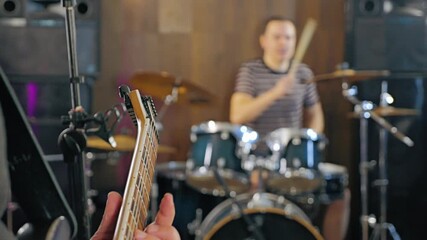 A guitarist plays in a studio, with focus on the guitar neck, while a drummer plays in the background. The ambiance suggests a casual jam session - Powered by Adobe