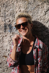 Portrait of cheerful woman in sunglasses and shirt standing near concrete wall