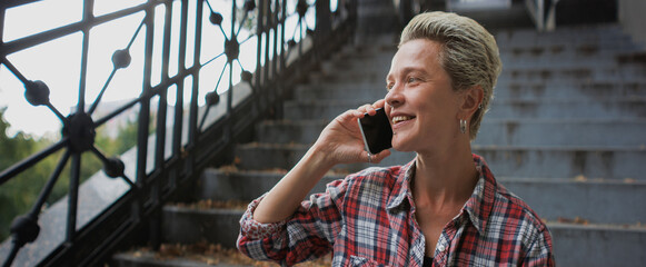 Positive short haired woman talking on smartphone on stairs in city, banner