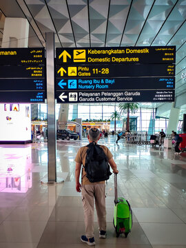 A man solo traveler with a backpack and a suitcase beside him faces and stares at the signboard inside Soekarno Hatta International Airport. Jakarta, Indonesia.