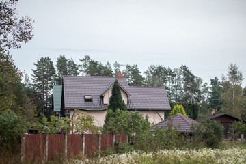 Fototapeta premium A beige suburban house with a dark tiled roof sits behind a wooden fence, surrounded by lush trees and vegetation under a cloudy sky.