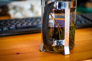 Close-up of fresh herbal tea leaves steeping in a French press, placed on a wooden desk near a computer keyboard.