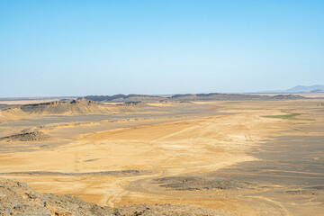 Vast Sahara desert plain in Morocco with golden sand patches dark rocky hills and distant horizon under clear blue sky creating wide panoramic view