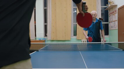 Rear view of player facing elderly man serving tennis ball on indoor table, awards arranged on shelf in background, focus on action and intensity as ball drops during competitive game